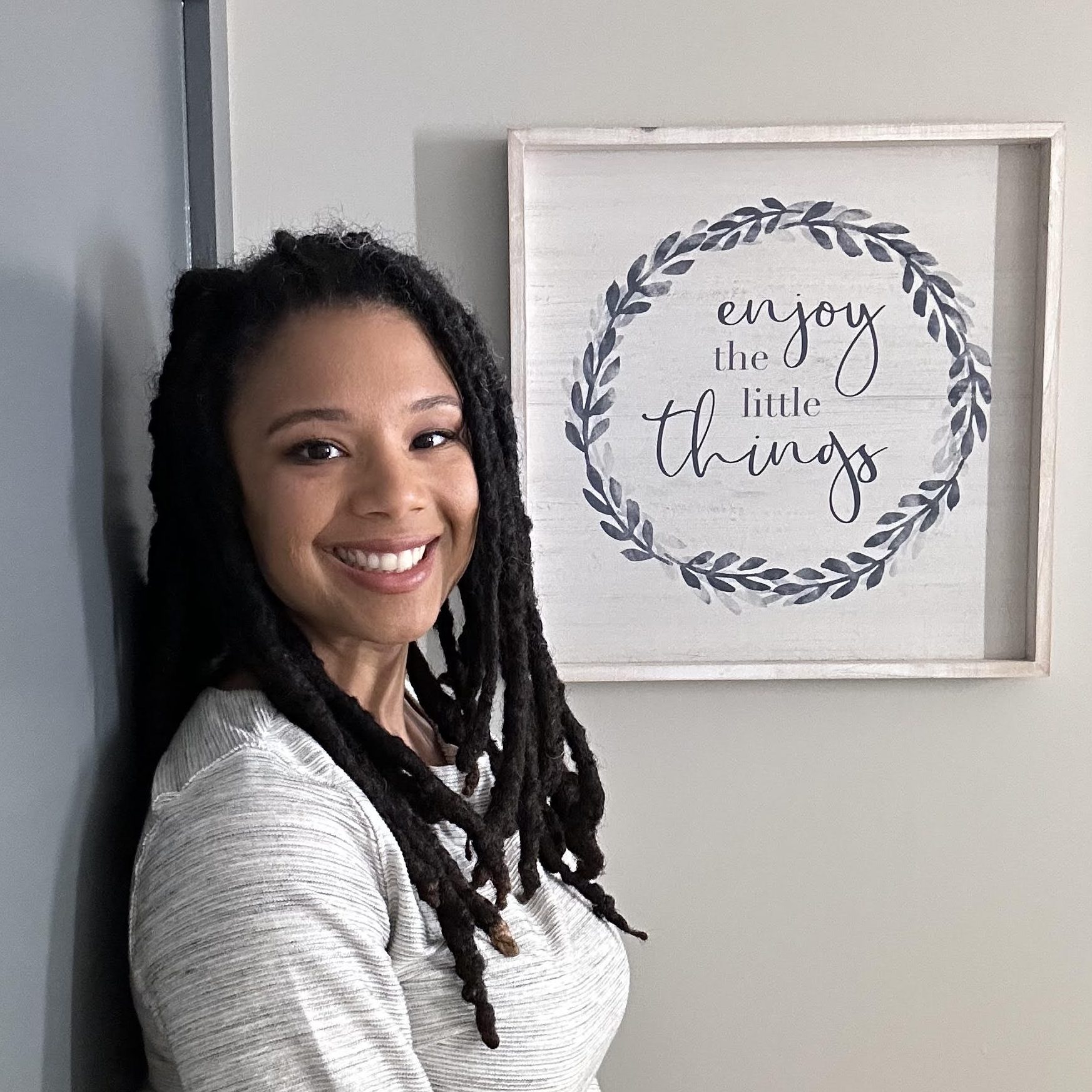 Portrait of Brittany smiling. She is an African American woman with long black locs wearing a white and gray striped dress. She is standing in front of a wooden picture that says, "Enjoy the Little Things". 